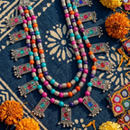 A vibrant, multi-strand beaded necklace with silver-toned geometric charms and colorful beads, styled on a dark blue patterned fabric with orange marigold flowers.
