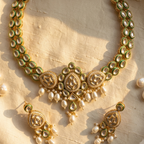 Gold-toned traditional jewelry set with a necklace and matching earrings, featuring green stone accents and white pearl-like drops, arranged on a textured beige fabric background with a few loose pearls nearby.