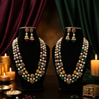 Two black jewelry display busts wearing ornate pearl and gemstone necklaces, with matching dangling earrings, arranged on a dark table with lit candles and draped burgundy and green fabric in the background.