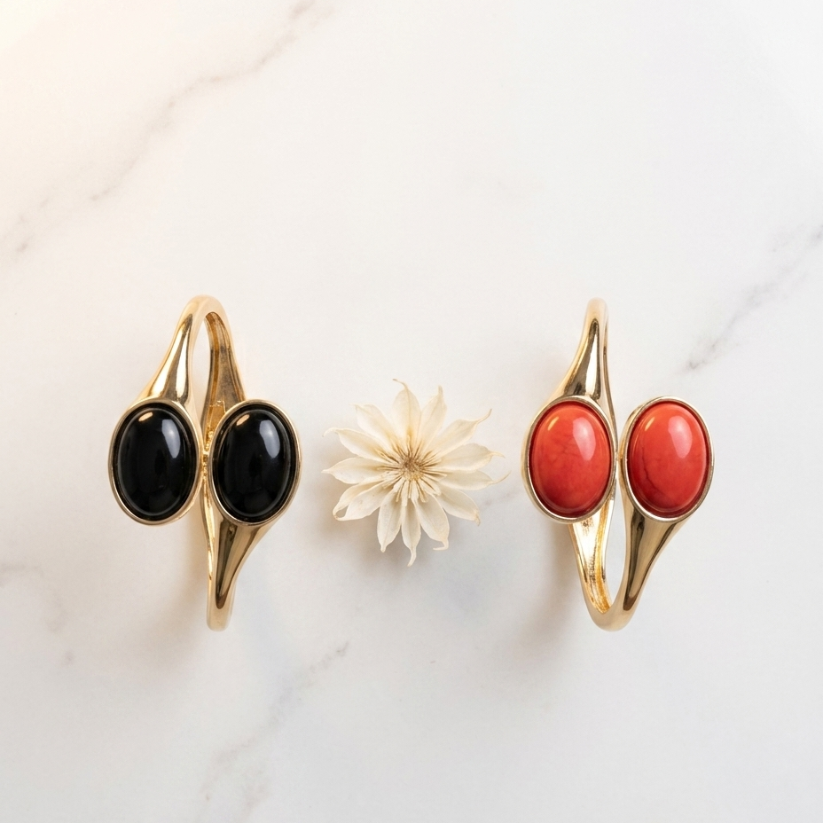 Two gold-toned wrap-style rings with oval cabochon stones, shown side by side on a white marble surface. The left ring has black stones, and the right ring has red-orange stones; a small white flower sits between them as a prop.