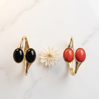 Two gold-toned wrap-style rings with oval cabochon stones, shown side by side on a white marble surface. The left ring has black stones, and the right ring has red-orange stones; a small white flower sits between them as a prop.