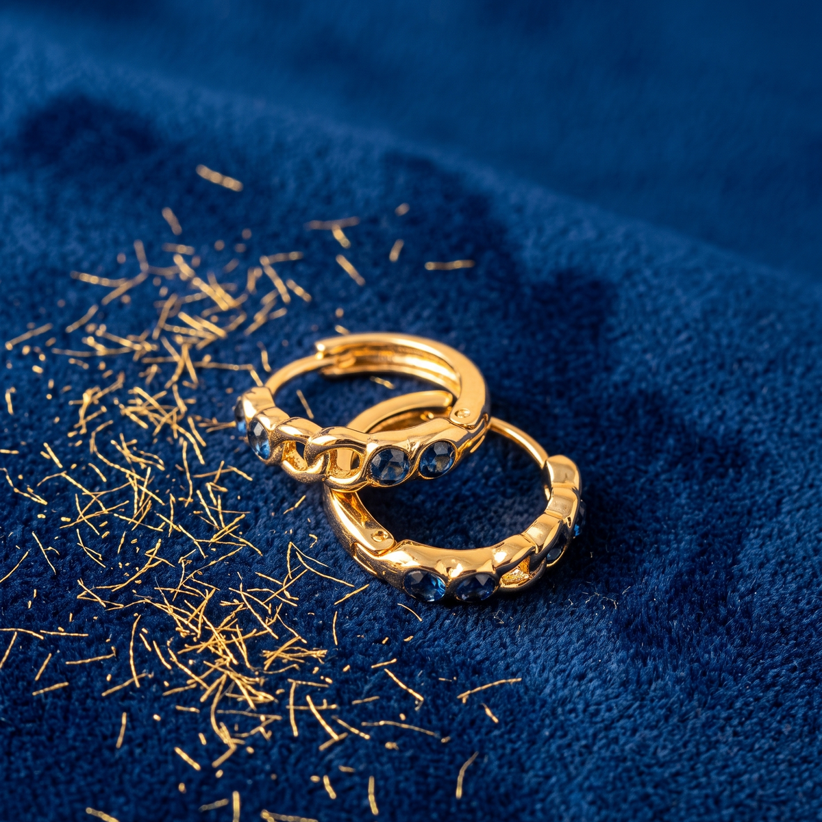 Two gold hoop rings with dark blue gemstones, resting on a textured navy blue fabric with scattered gold shavings.