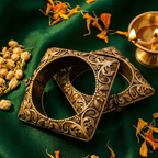 Two ornate antique-style bronze rings with intricate floral engravings, displayed on a dark green satin fabric with dried orange flowers and a brass diya in the background.