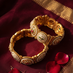 Two ornate gold bangles with white enamel inlays and red gemstone accents, arranged on a deep maroon fabric with gold border trim and scattered red rose petals.
