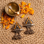 Two ornate silver-toned Indian jhumka earrings with orange stone accents and dangling bell charms, arranged on a woven straw mat with scattered orange flower petals and a brass diya in the background.