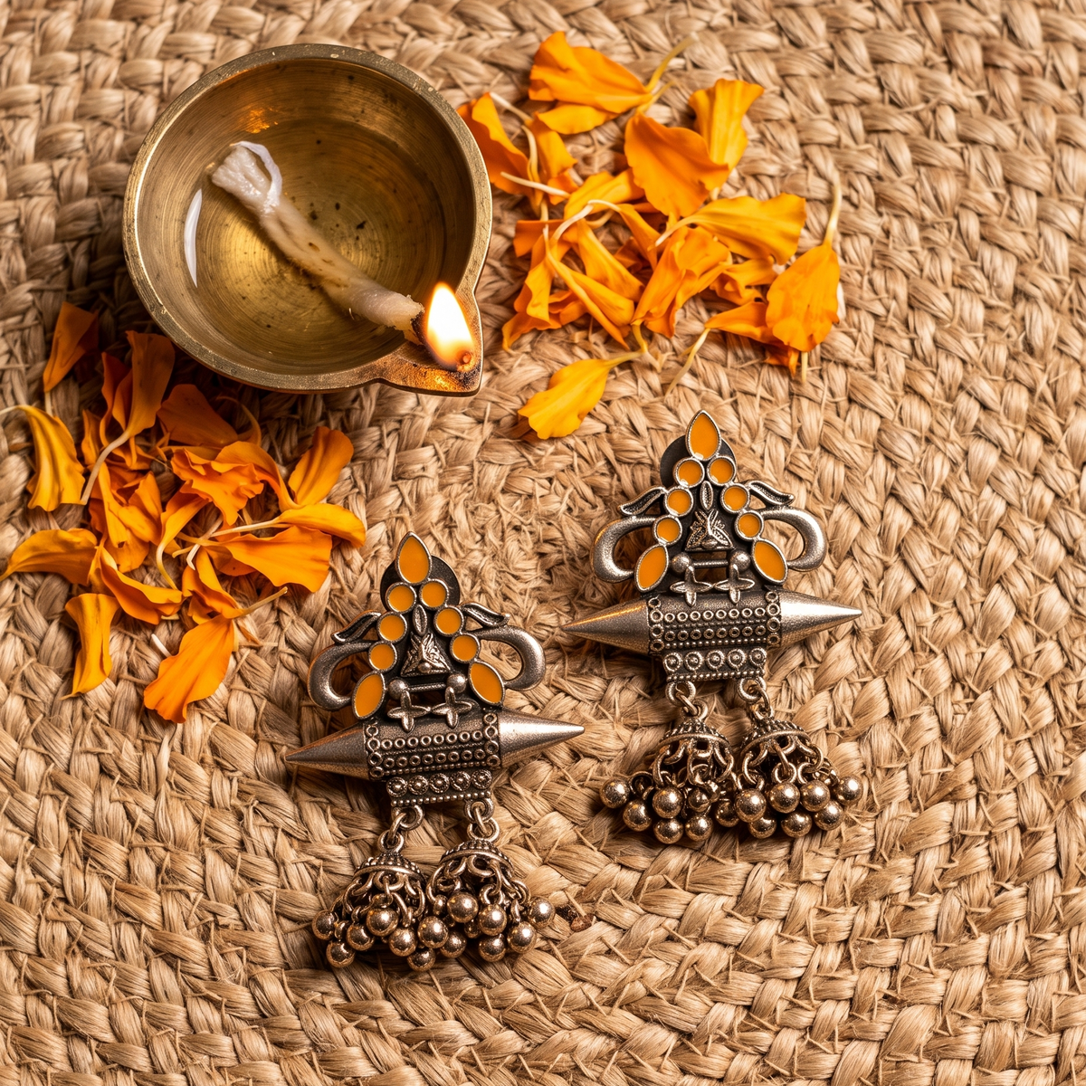 Two ornate silver-toned Indian jhumka earrings with orange stone accents and dangling bell charms, arranged on a woven straw mat with scattered orange flower petals and a brass diya in the background.