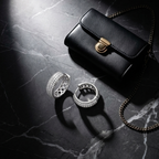 Two silver rings with diamond-like embellishments are displayed on a dark marble surface next to a small black leather handbag with a gold clasp and chain strap, under dramatic spotlighting.