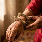 A close-up of a person's hands adorned with intricate brown henna designs, wearing ornate gold-toned traditional Indian wrist cuffs (kada) over a red embroidered sleeve. The background is a softly blurred warm indoor setting.