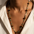 A close-up of a person wearing a long beaded necklace with green beads, tiger's eye beads, and clear rectangular beads over a white shirt.