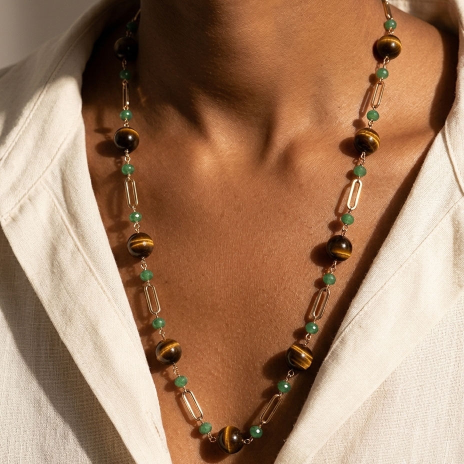 A close-up of a person wearing a long beaded necklace with green beads, tiger's eye beads, and clear rectangular beads over a white shirt.