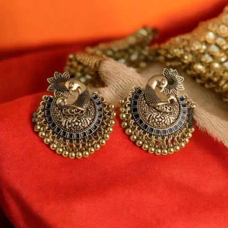 Yugavi Arin antique gold peacock crescent stud earrings displayed as a pair on white background — full Mayura bird form with engraved flower crest at crown, densely worked dotwork and scrollwork gold body, row of square navy blue enamel tiles along inner crescent arc, and cascading gold ghungroo ball fringe drops along the base