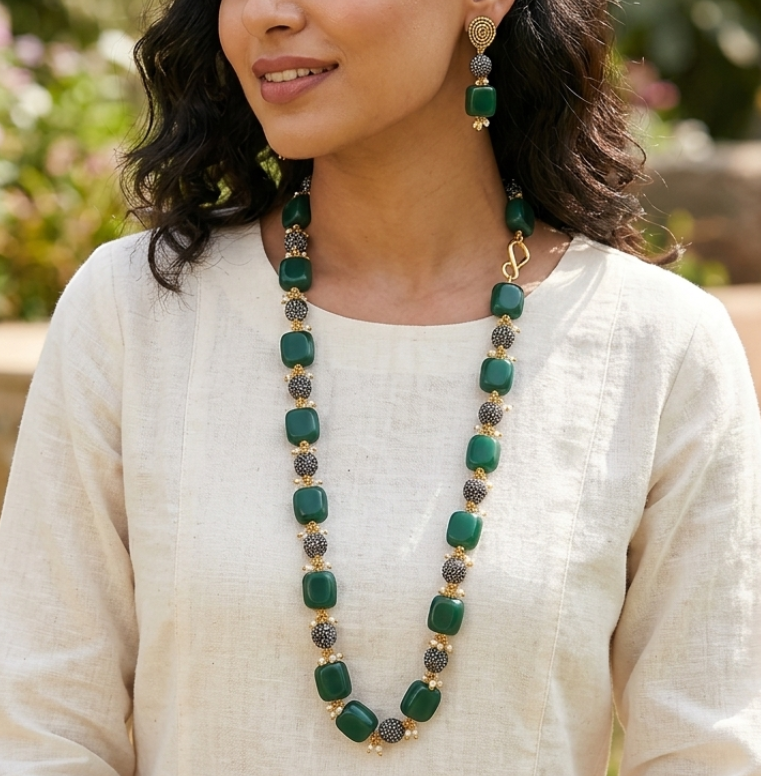 Woman wearing a long green necklace and earrings outdoors with blurred greenery in the background