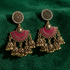 Yugavi Devanshi antique gold triple jhumka earrings displayed as a pair on hot pink magenta background — large concentric-ring gold disc stud tops, deep magenta enamel half-moon arch connectors with geometric gold border and centre dome, three cascading antique gold jhumka bells with bead drops hanging below each arch