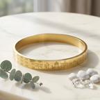 Gold bracelet with engraved text on a marble surface with eucalyptus leaves and stones.