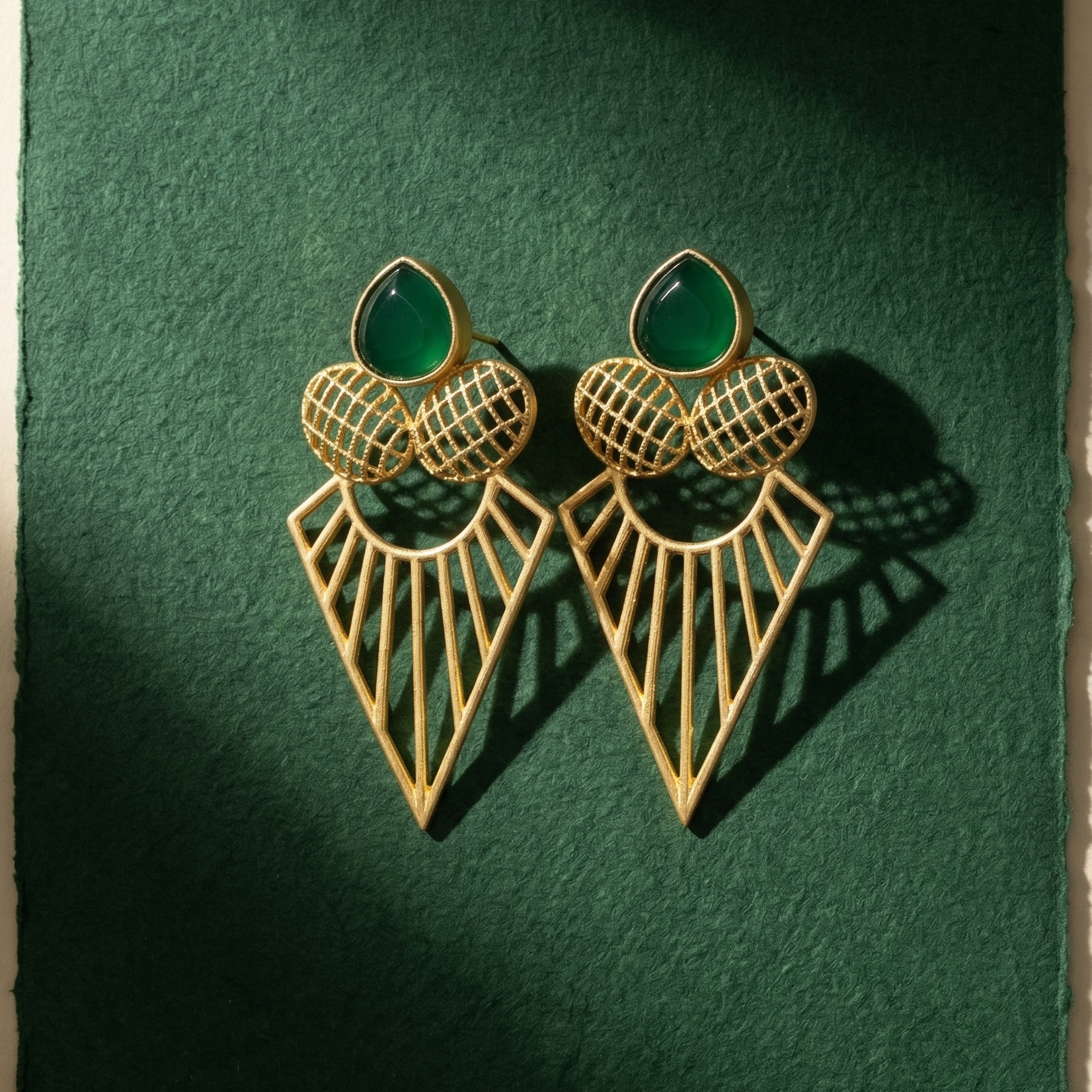 Yugavi Kirana geometric gold drop earrings displayed as a pair — emerald green teardrop cabochon stud with two cross-hatched grid gold ovals, and a large open-work triangular pendant with radiating sun-ray gold lines, gold polish finish on khaki fabric