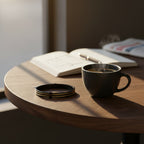 Black coffee cup with steam on a wooden table next to an open book and a newspaper.