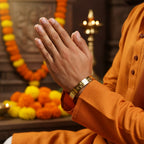 Person in traditional attire with hands clasped in a gesture of prayer or respect, with decorative elements in the background.