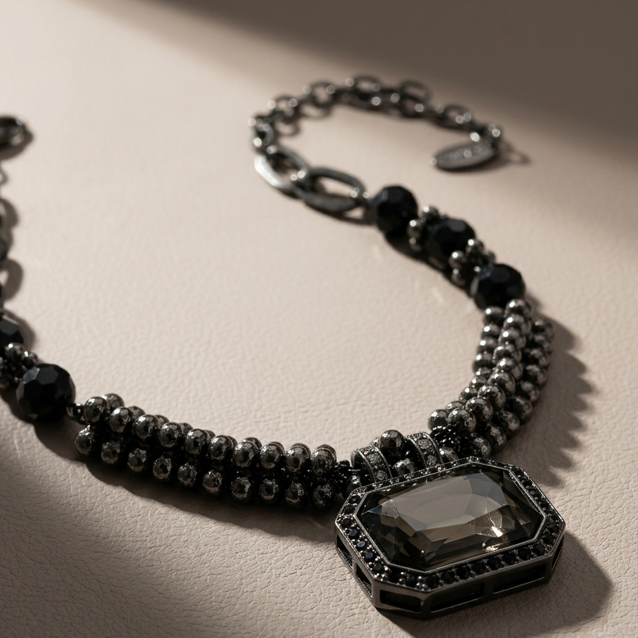 A close-up of a black beaded necklace with a faceted smoky-gray octagonal pendant, styled on a textured beige surface with dramatic diagonal shadows.
