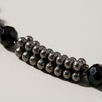 A close-up of a beaded strand featuring rows of metallic gunmetal gray beads flanked by faceted black end beads, resting on a neutral light gray surface.
