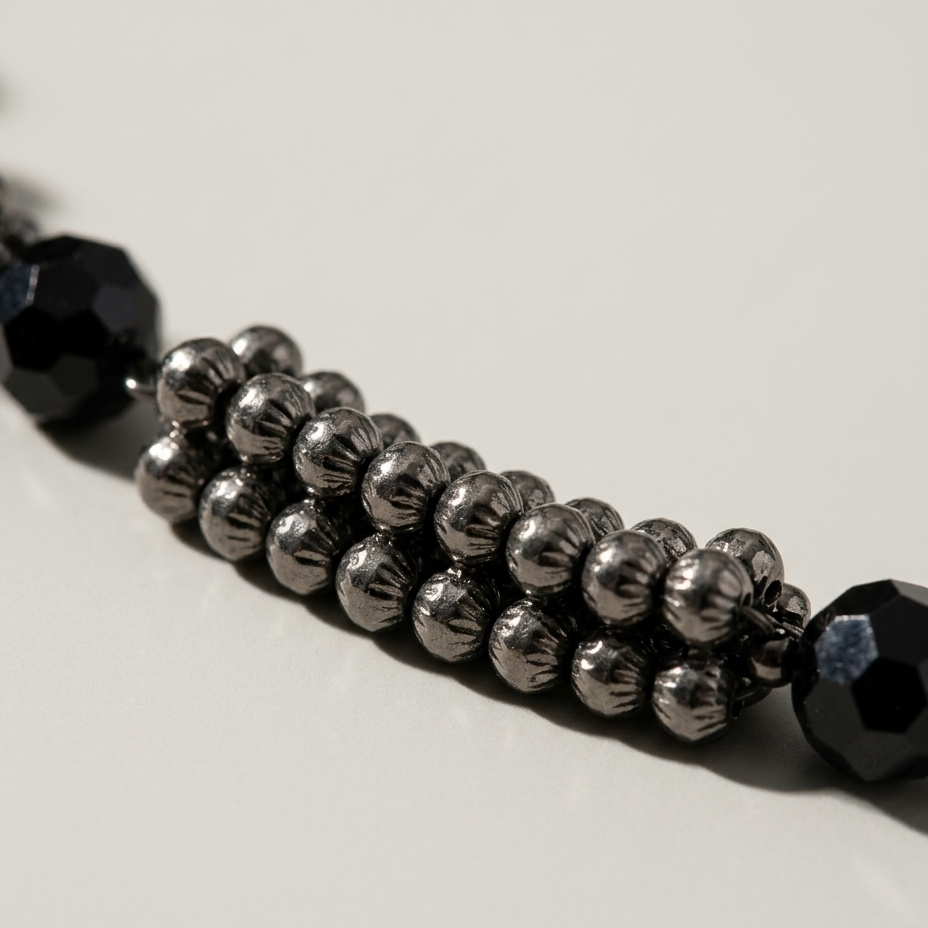 A close-up of a beaded strand featuring rows of metallic gunmetal gray beads flanked by faceted black end beads, resting on a neutral light gray surface.