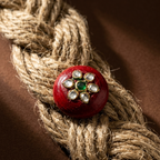A round red decorative button or brooch with a floral gold-tone setting, white stones, and a central green stone, resting on a thick braided beige rope against a dark brown background.