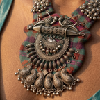 A close-up of a person wearing an ornate silver-toned statement necklace with paisley and floral motifs over a colorful patterned fabric bib, paired with a teal top.
