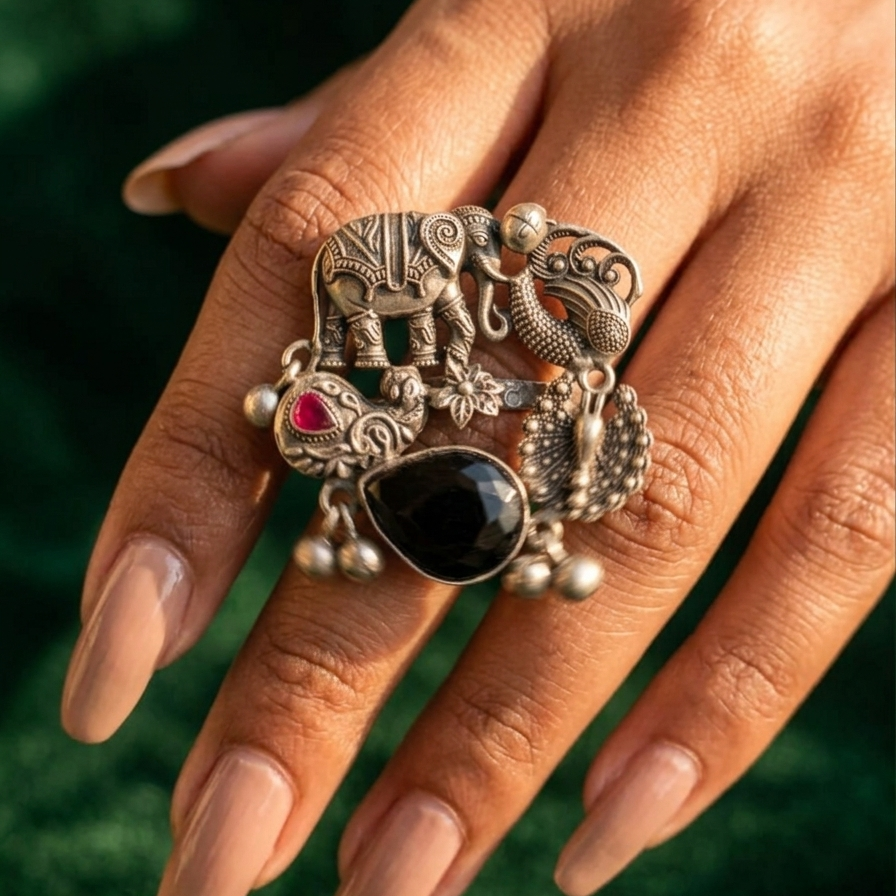Yugavi Gajendra oxidised antique silver statement ring — clearly showing carved elephant in temple caparison standing beside a textured peacock on band face, with three hanging drops: large black teardrop stone, ruby red accent and peacock fan form with silver balls below