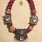 A traditional Indian statement necklace with a deep magenta braided cord, adorned with ornate silver and gold-plated metal plates, white stones, and small pearl bead drops, displayed on a textured beige background.