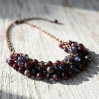 A beaded collar necklace with faceted dark red, navy blue, and smoky gray beads on a copper-toned chain, displayed on a weathered white wooden surface.